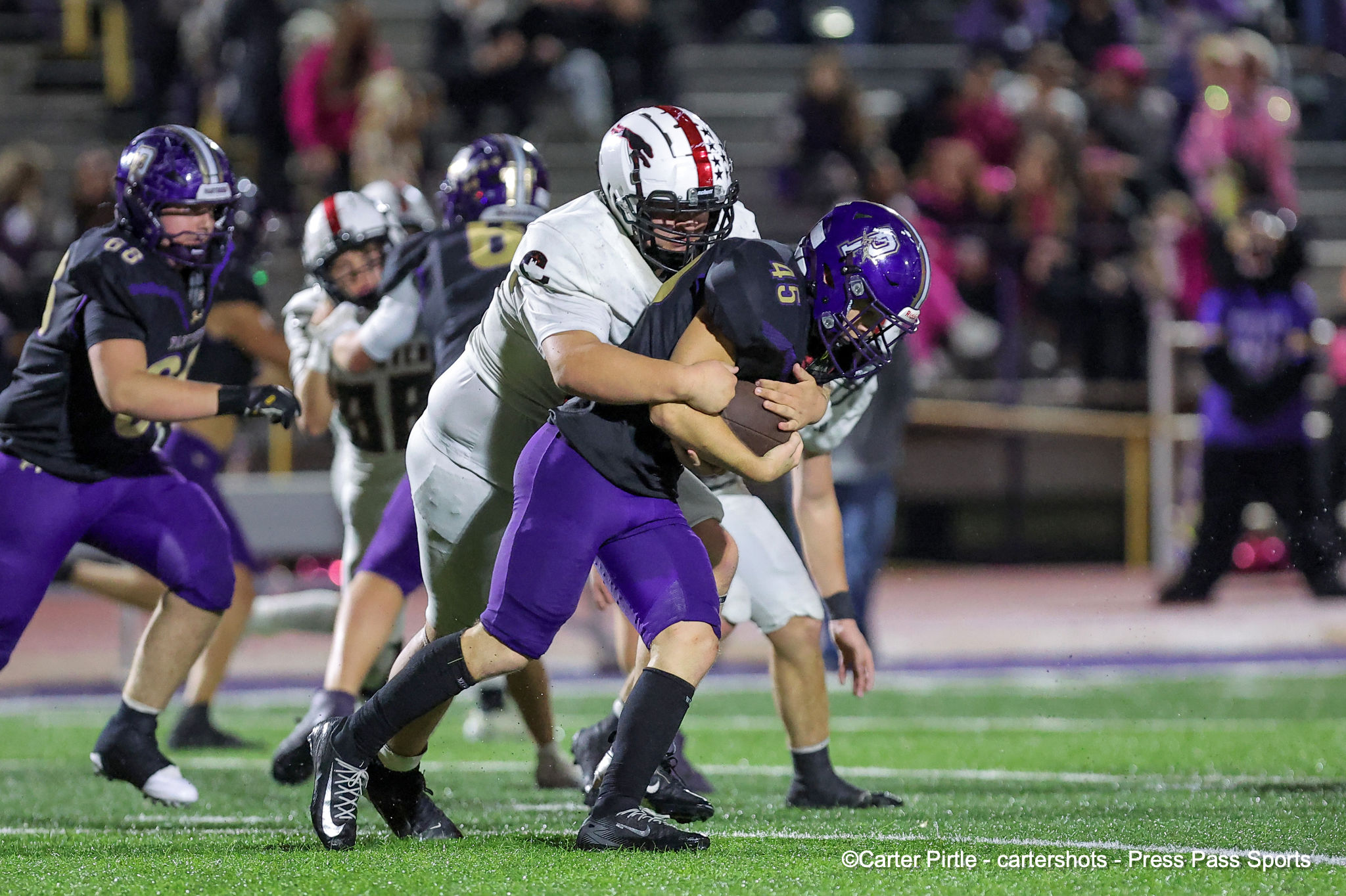 Two football players in purple and white collide for the ball on a green turf during a night game, with a blurred crowd in the stands behind them.