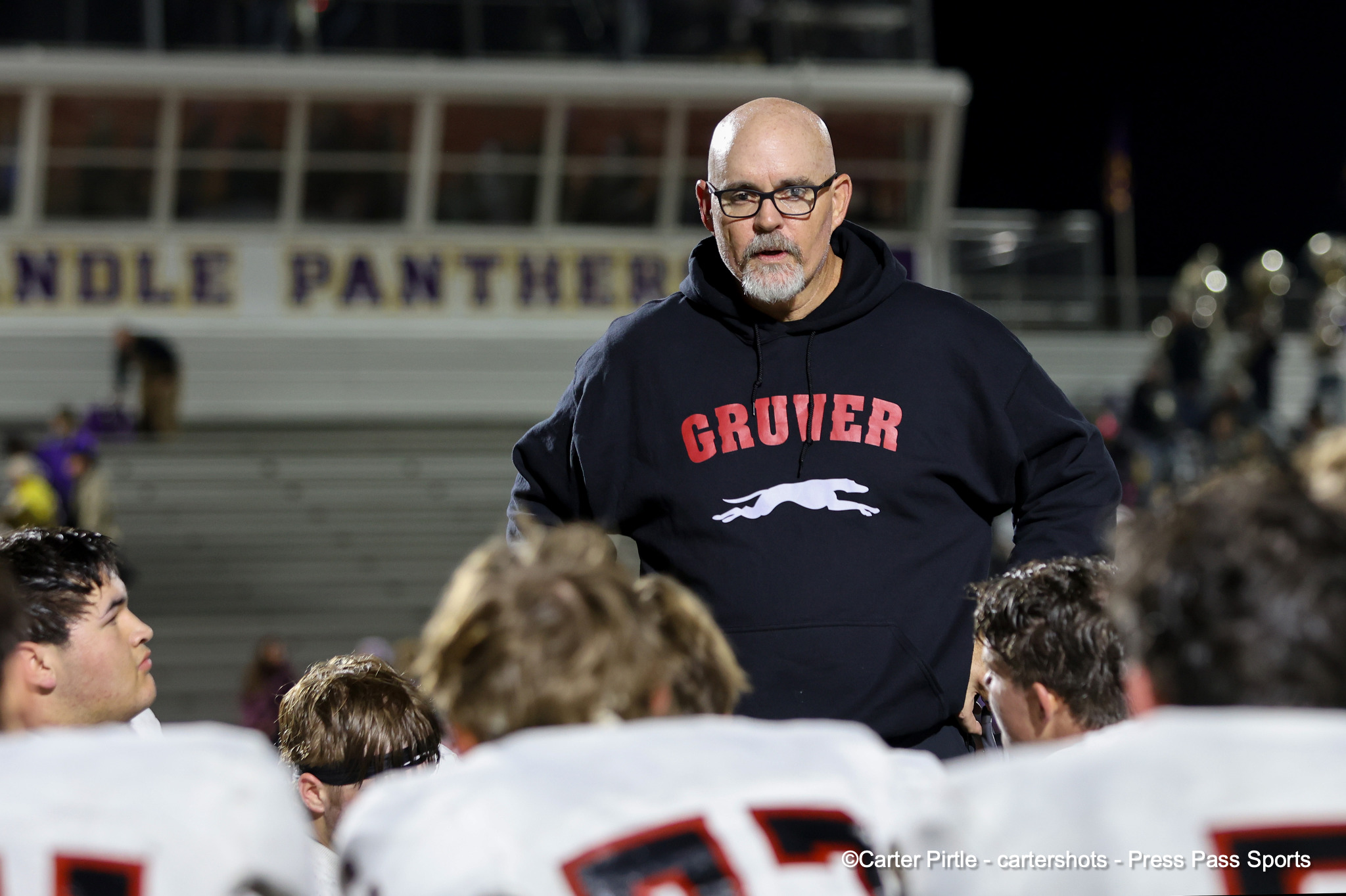 Man with glasses and a gray beard wearing a dark hoodie, standing in front of a crowd at a sports stadium.