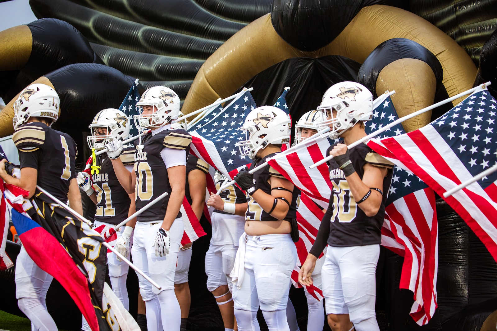 Football players in black and gold uniforms march with American flags under a large inflatable tunnel wall; a pregame entry scene.