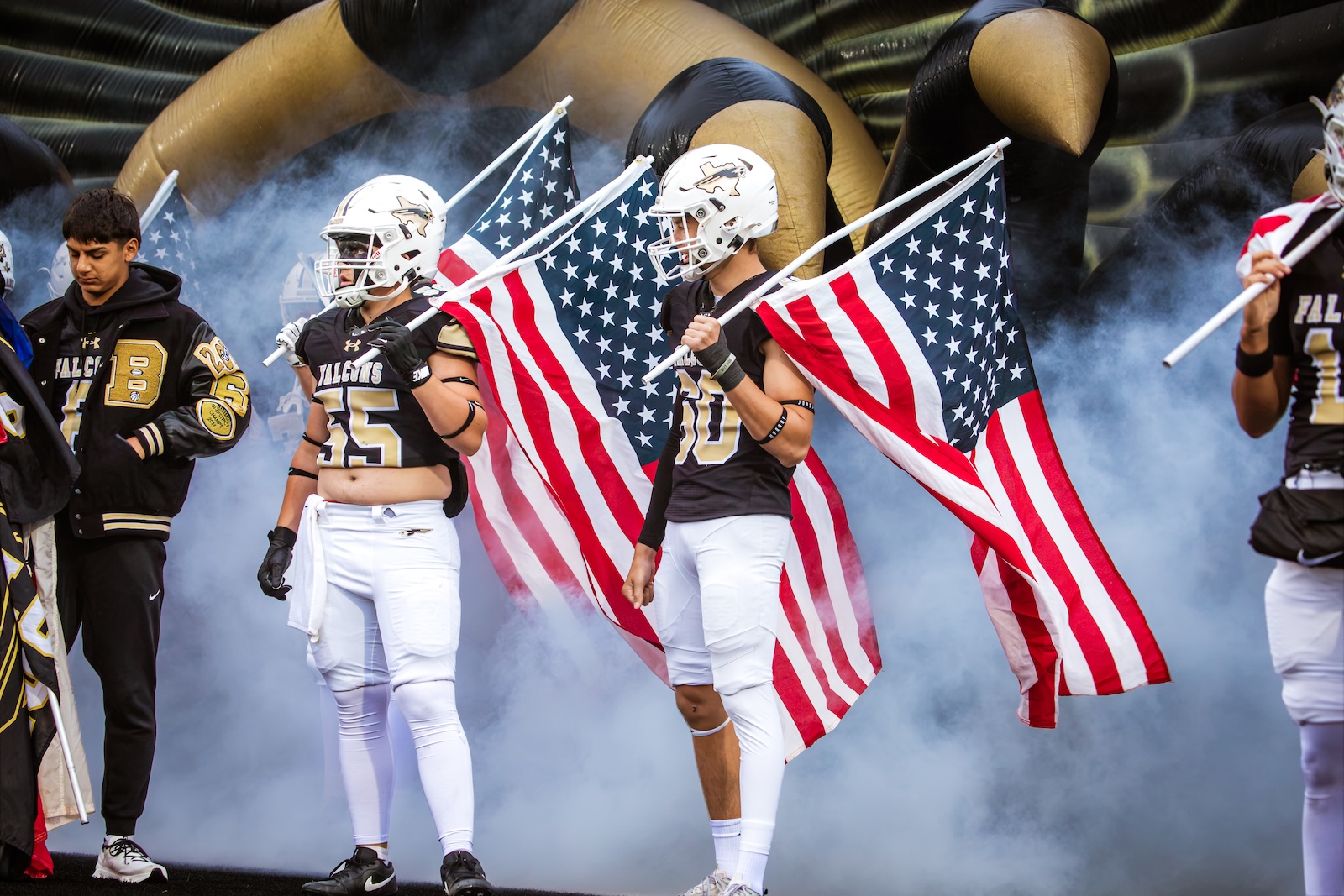 Football players in black and gold uniforms carry American flags through a smoky stadium entrance.