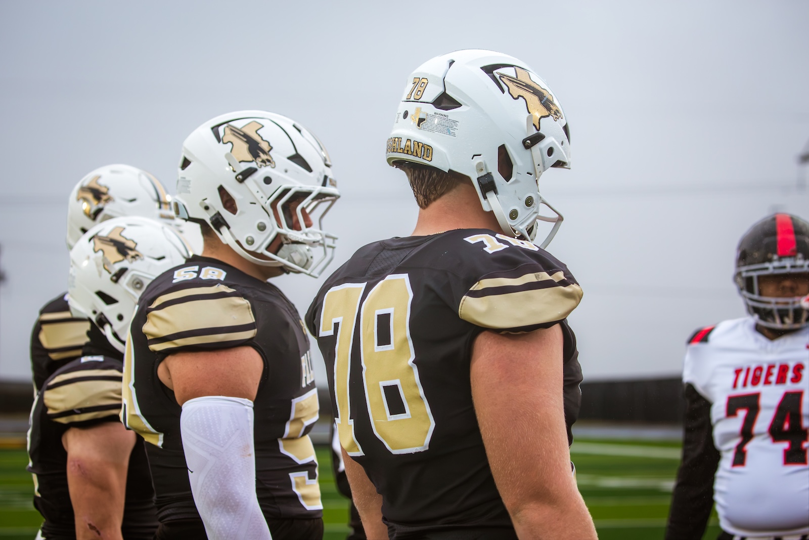 Line of football players in black and gold uniforms standing on a field, wearing white helmets with a gold logo.