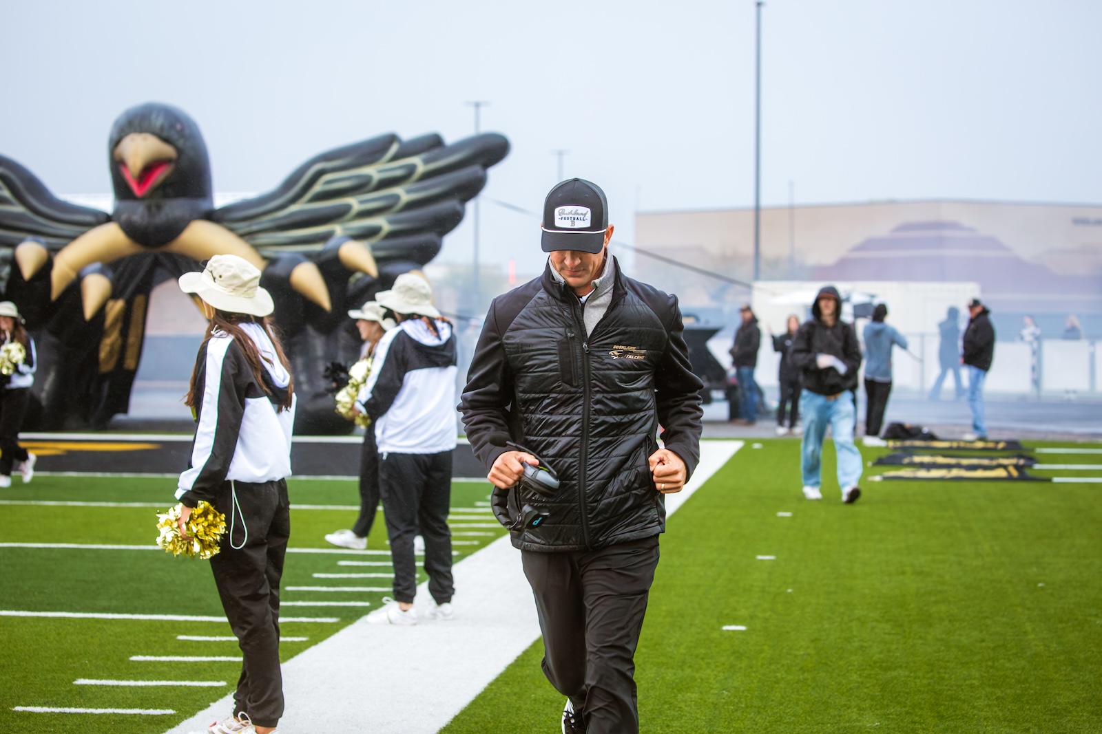 Man in a black puffer jacket walking on a football field, with cheerleaders and a large inflatable eagle in the background.