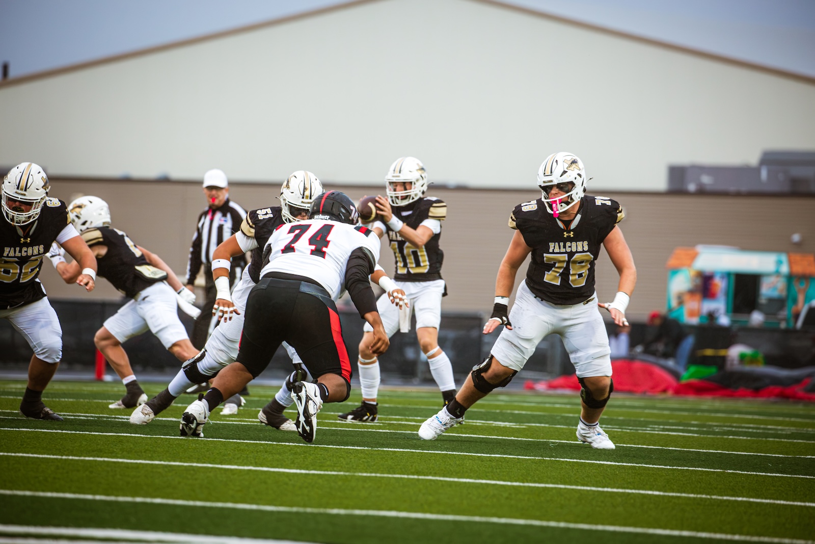 American football game scene: Falcons players in black and gold jerseys clash on a green turf as a defender in white (#74) linemen up against their blockers near the line of scrimmage.