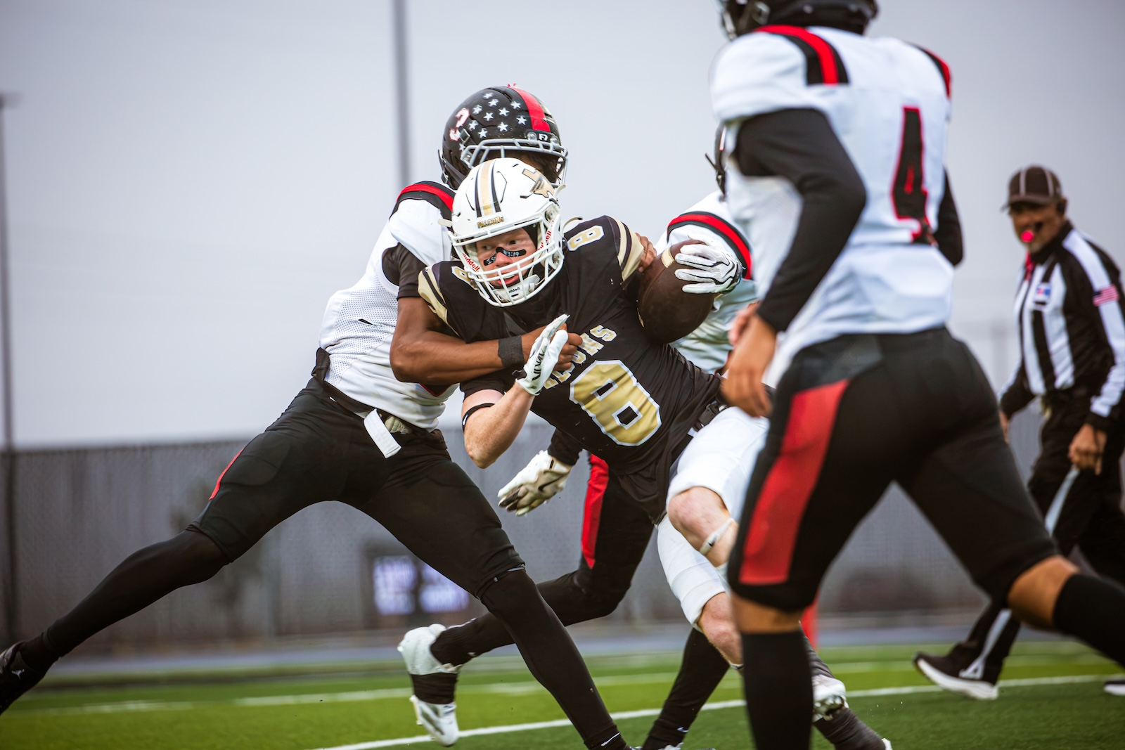 Action shot of a running back in black and gold being tackled by a defender in white, on a football field with a chain-link fence in the background.