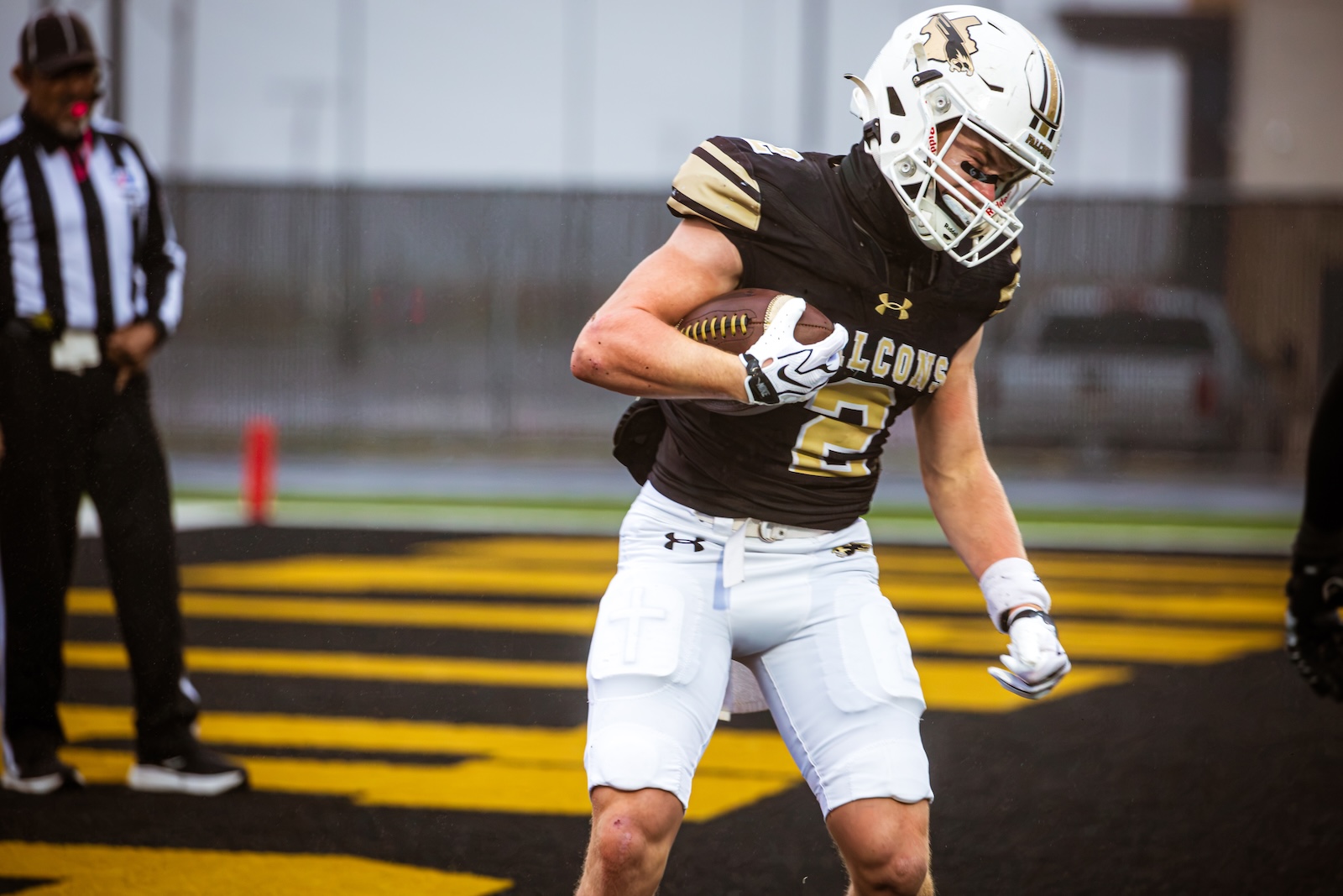 American football player in black and gold Falcons uniform runs with the ball into the end zone, referee watching in the background.