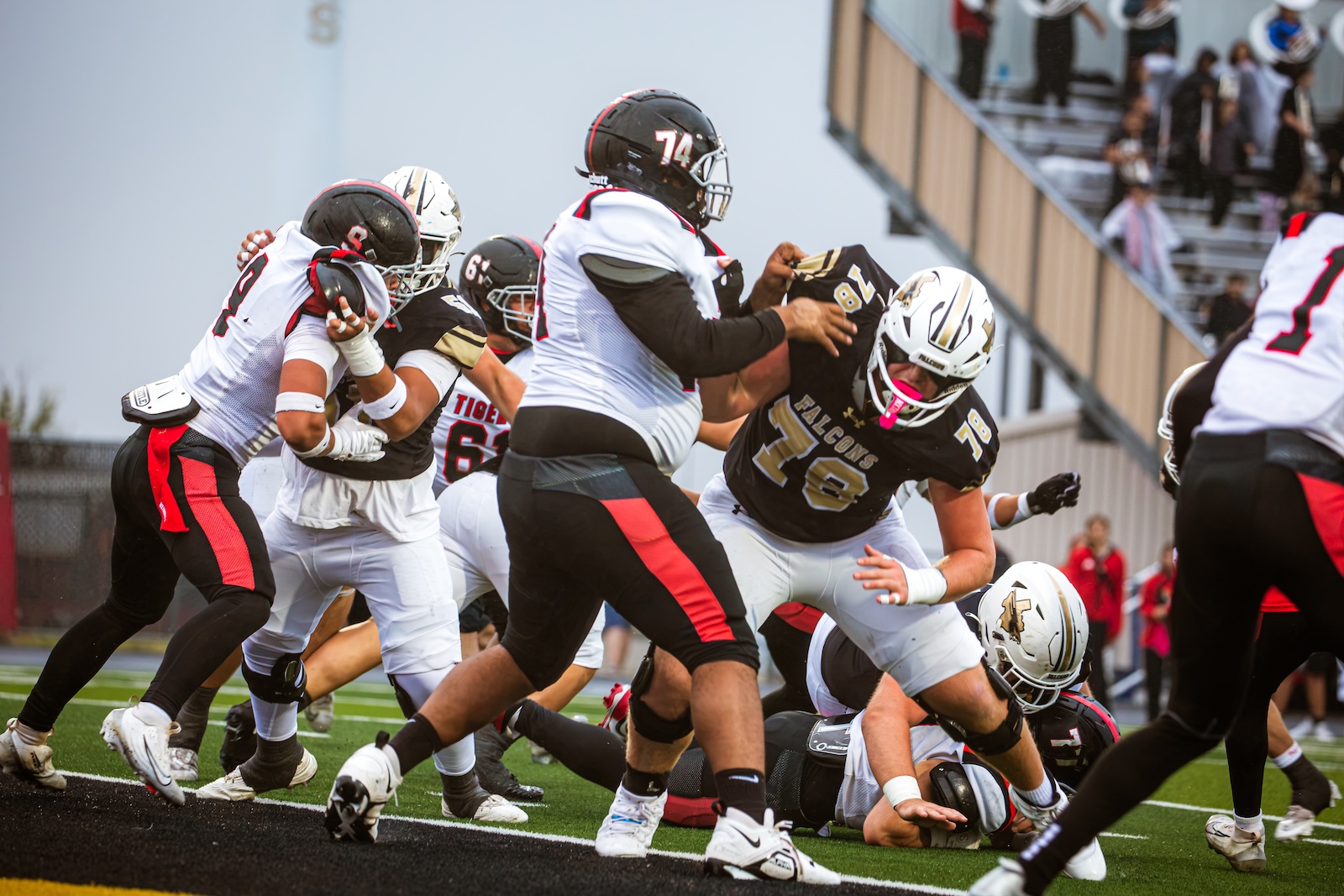 Action-packed football scene with players in white and black uniforms colliding near the ground on a turf field, crowd in stands in the background.