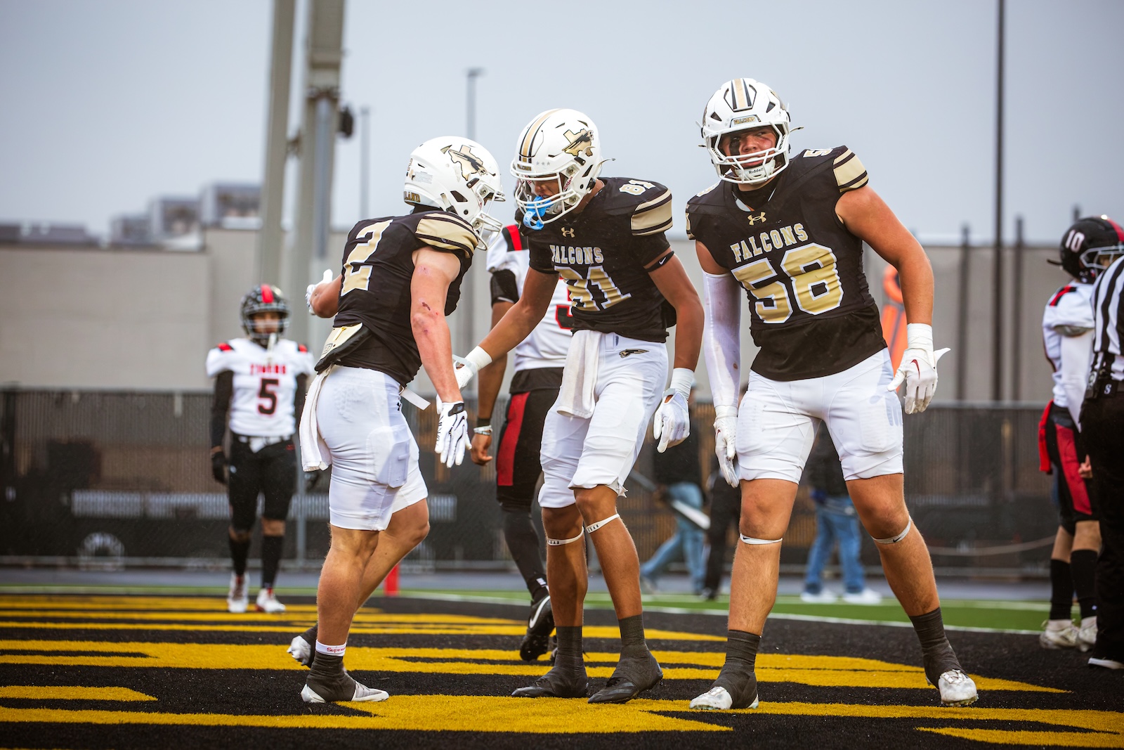 Three Falcons football players wearing black and gold jerseys celebrate together on a yellow-and-black end zone field.