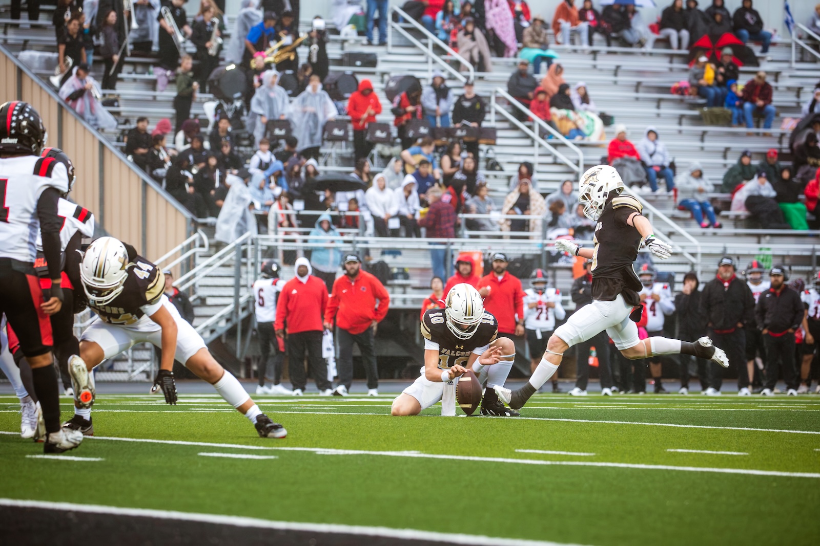 Two football players in black and white uniforms on a green turf; one kneels to place the ball as another player leaps in the air, with a crowded stadium in the background.