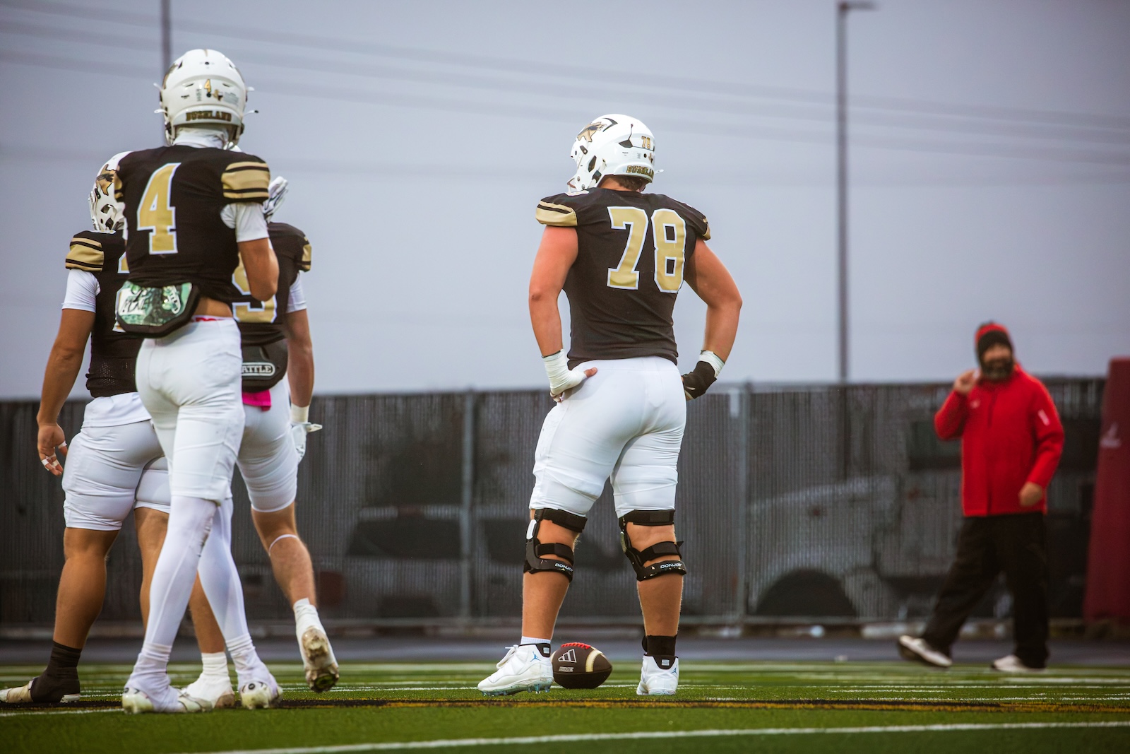 American football players in black and gold uniforms stand on a green turf field during practice, with the center foregrounded wearing jersey number 28 and knee braces.
