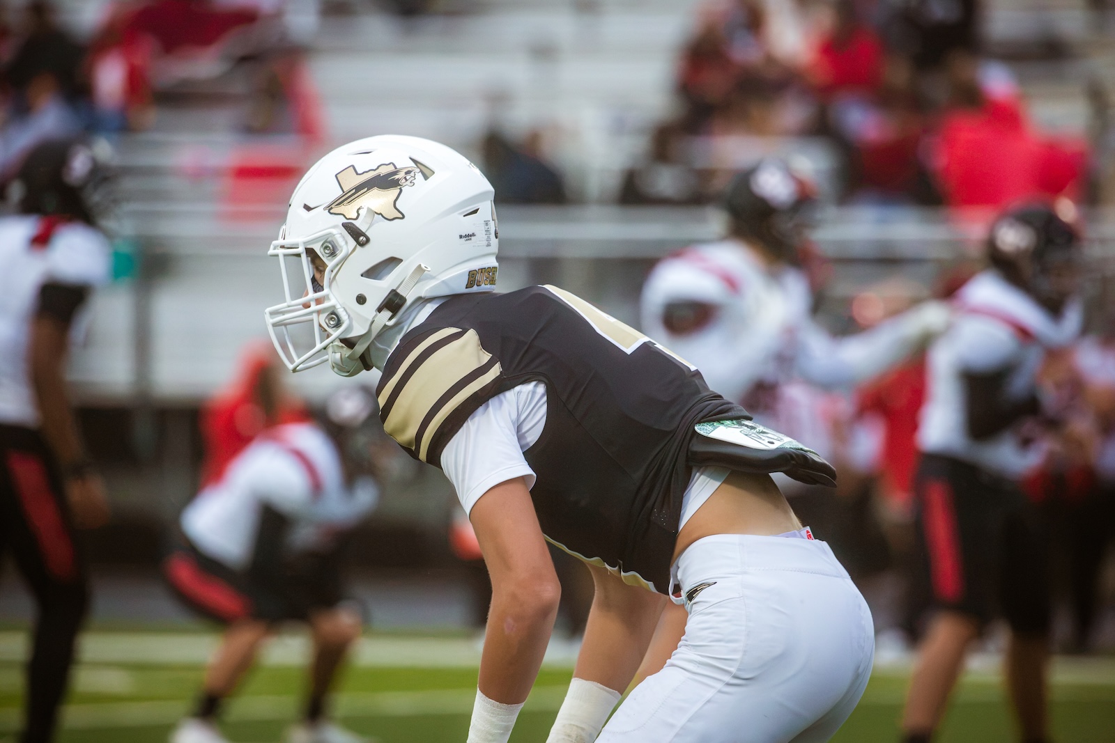 Football player in a white helmet and black and gold uniform crouches on the field during a game with blurred stands in the background.