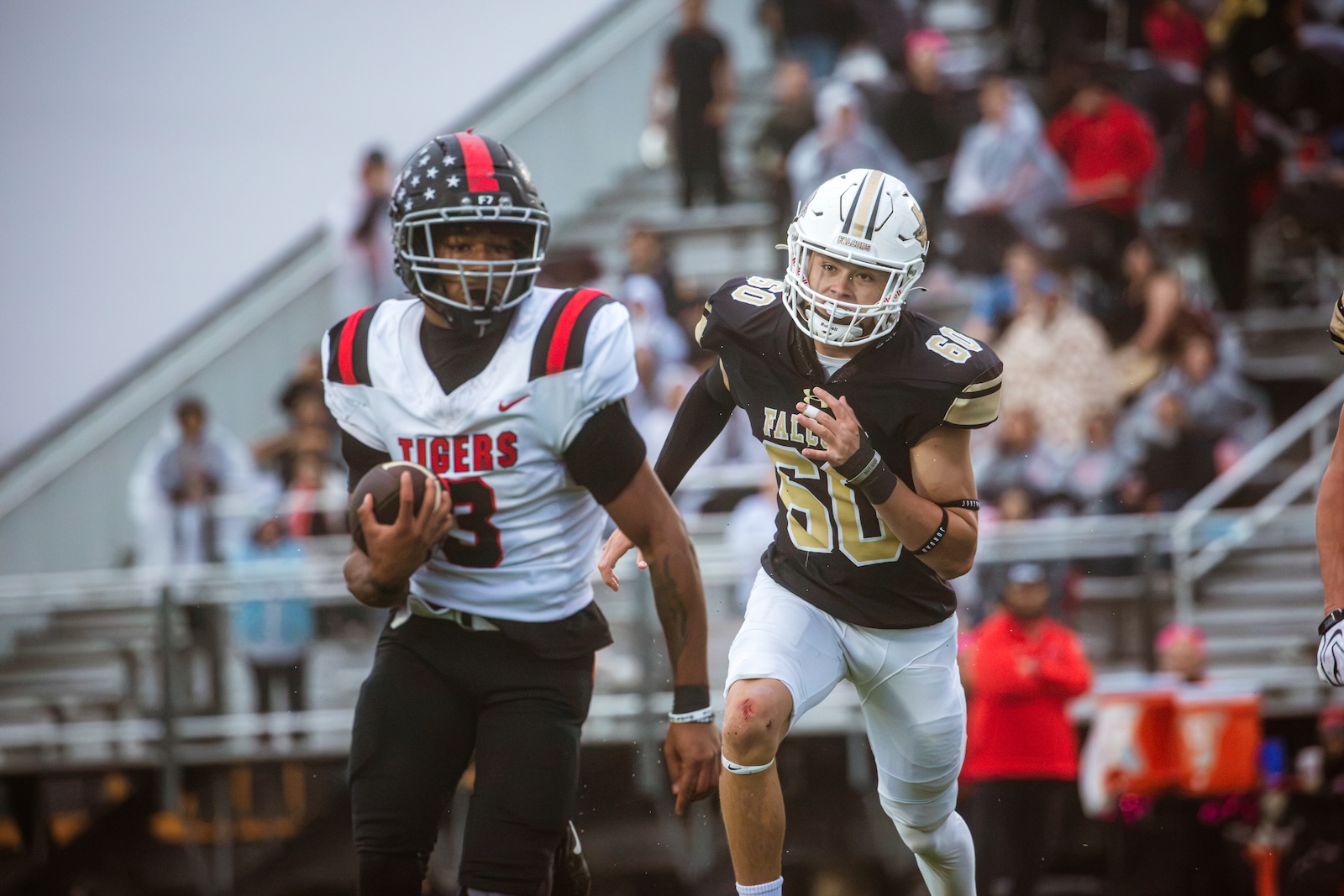 Tigers ball carrier in a white jersey runs with the football, chased by a Falcons defender in black; blurred spectators in the stands behind them.
