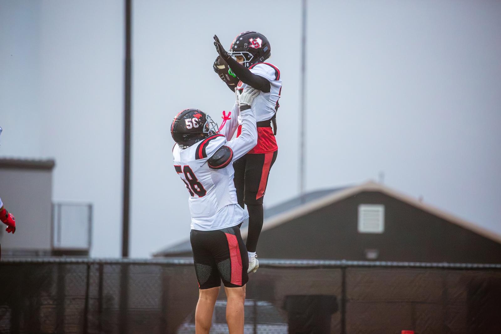 Two football players in white and red jerseys jump, one lifting the other to catch a pass against a chain-link fence.