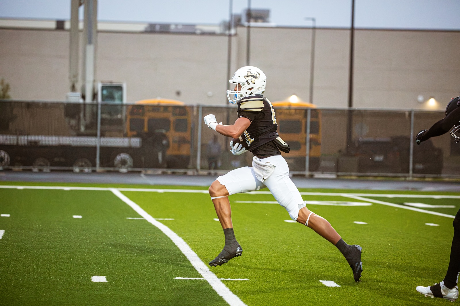 Football player in black and gold uniform running on a green turf field, holding a ball and wearing a white helmet, with outstretched arms and clear motion.