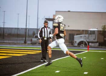 _V4A7545 | Press Pass Sports Football player in a black-and-gold uniform (#81) leaps toward the end zone as a referee watches from the sideline on a turf field.