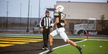Football player in a black-and-gold uniform (#81) leaps toward the end zone as a referee watches from the sideline on a turf field.