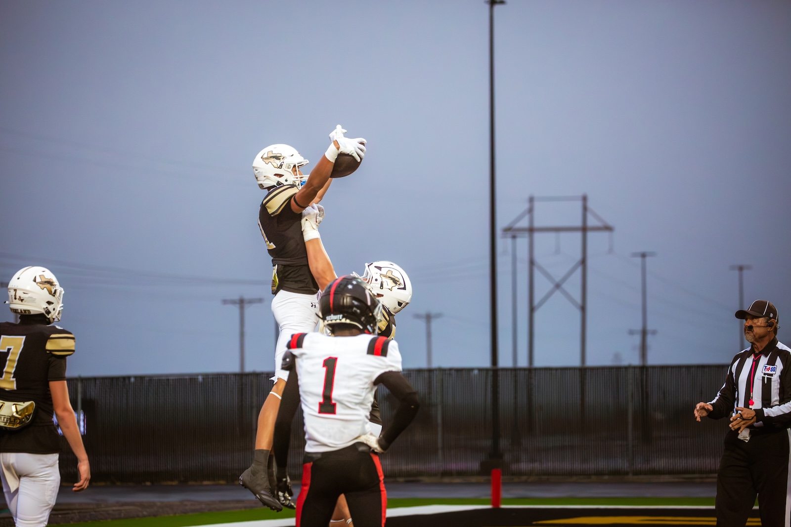 Football players reach for a high-spirited catch as a defender leaps to reel in the ball near the end zone with a referee watching from the right.