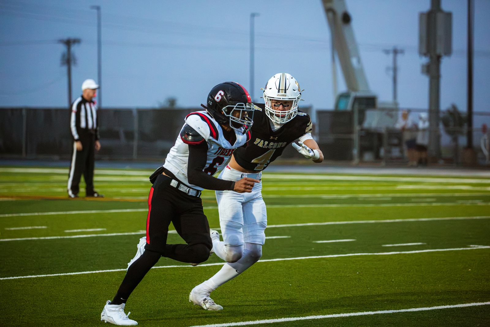 Two football players race down the field in foreground—one in a black Falcons uniform, the other in white—while a referee watches in the background on a green turf field.