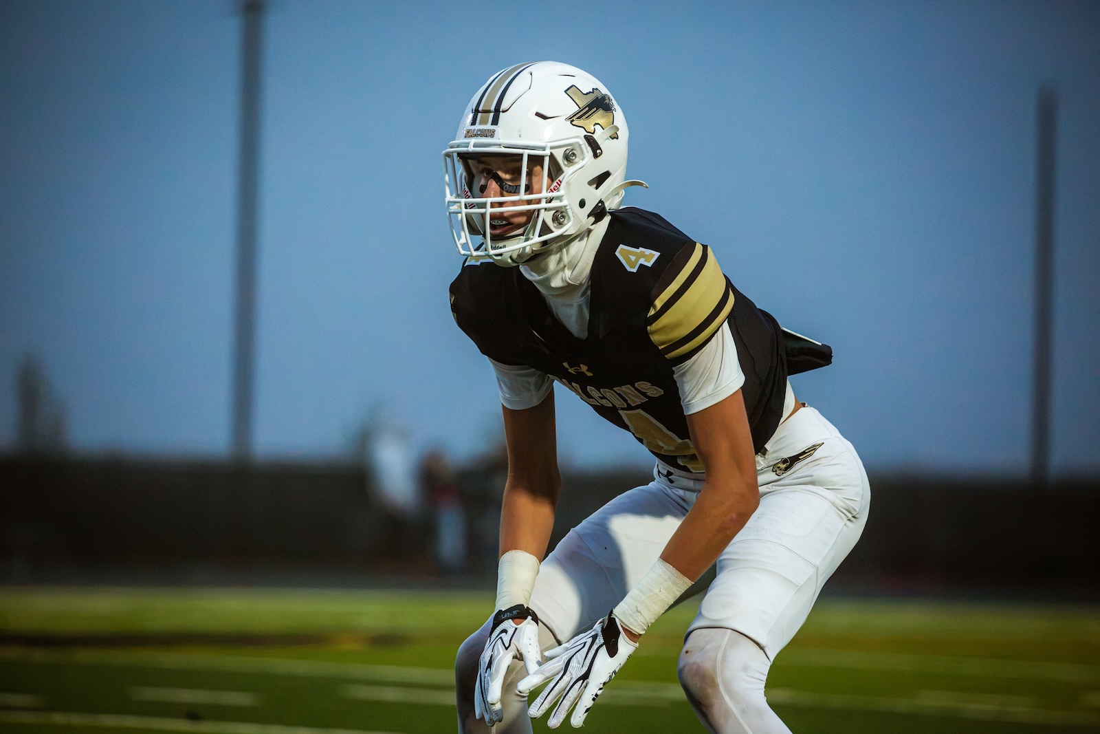 American football player in a black and gold uniform crouches in a ready stance on the field, wearing a white helmet with a face mask.