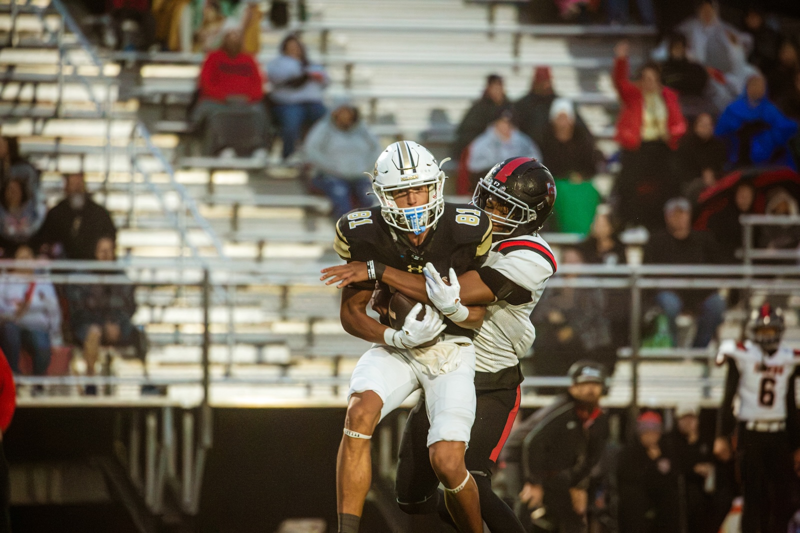 Two football players collide on the field during a game; one in a black/gold uniform holds the ball as a defender in white attempts a tackle, with blurred spectators in the stands behind.