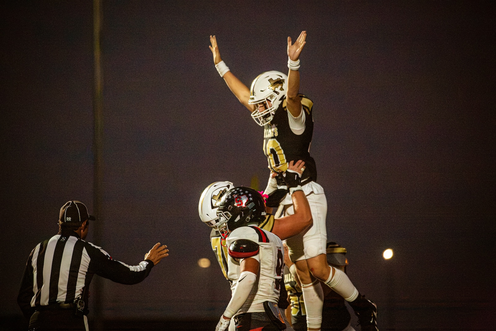 Football players celebrate on the field at night, lifting a teammate onto their shoulders.