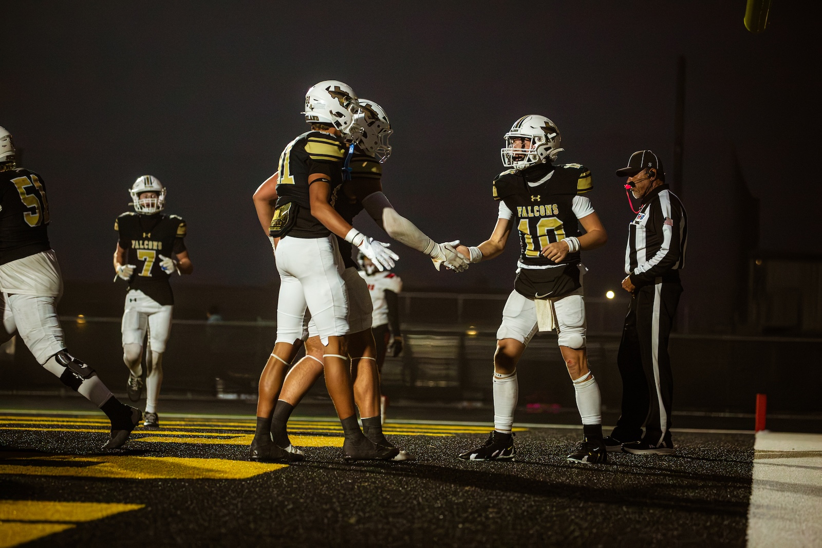 Football players in black and gold Falcons uniforms high-fiving on a wet night field with a referee nearby.