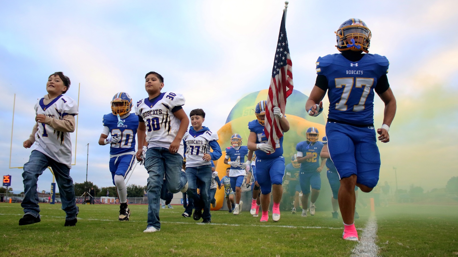 Young football players run onto the field from a colorful inflatable tunnel, one carrying the American flag, wide in blue and white uniforms.