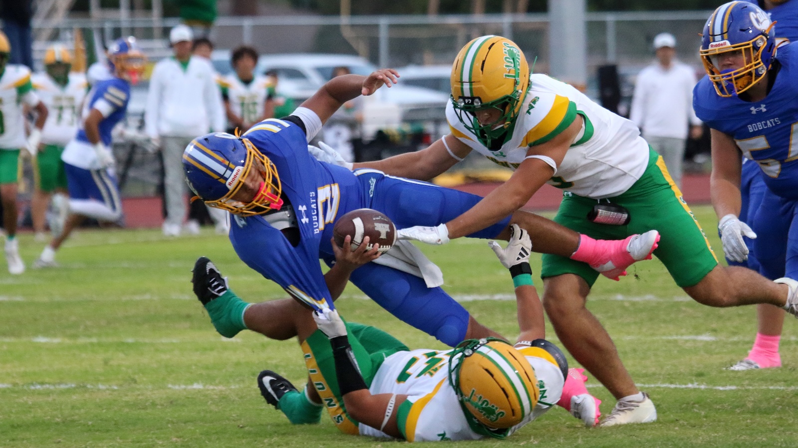 Football players in blue and green chase a ball; a blue team runner dives while being tackled by a green teammate on a grassy field.