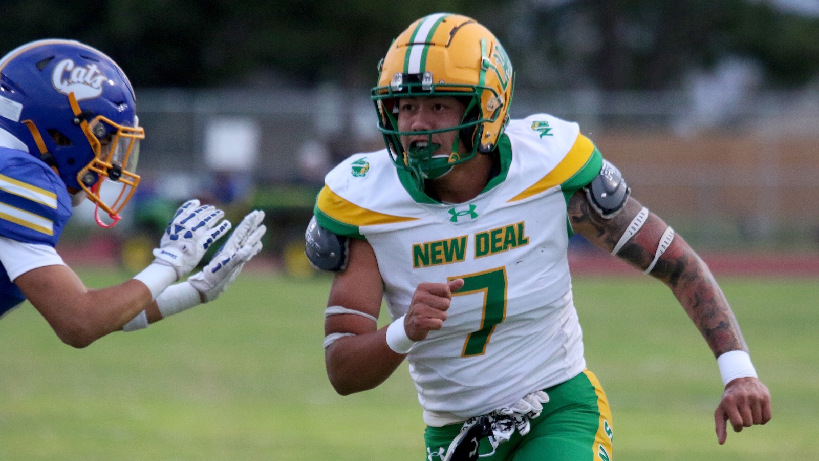 Football player in a white and green New Deal jersey (#7) runs with the ball as a blue-clad defender closes in on a grass field.