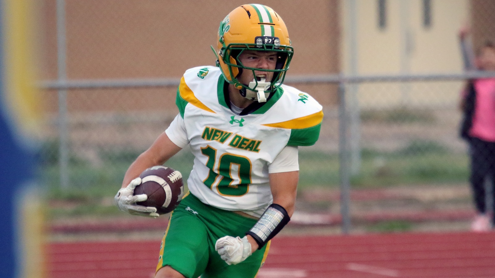 Football player in a white and green New Deal jersey, number 10, running with the ball on a track field.