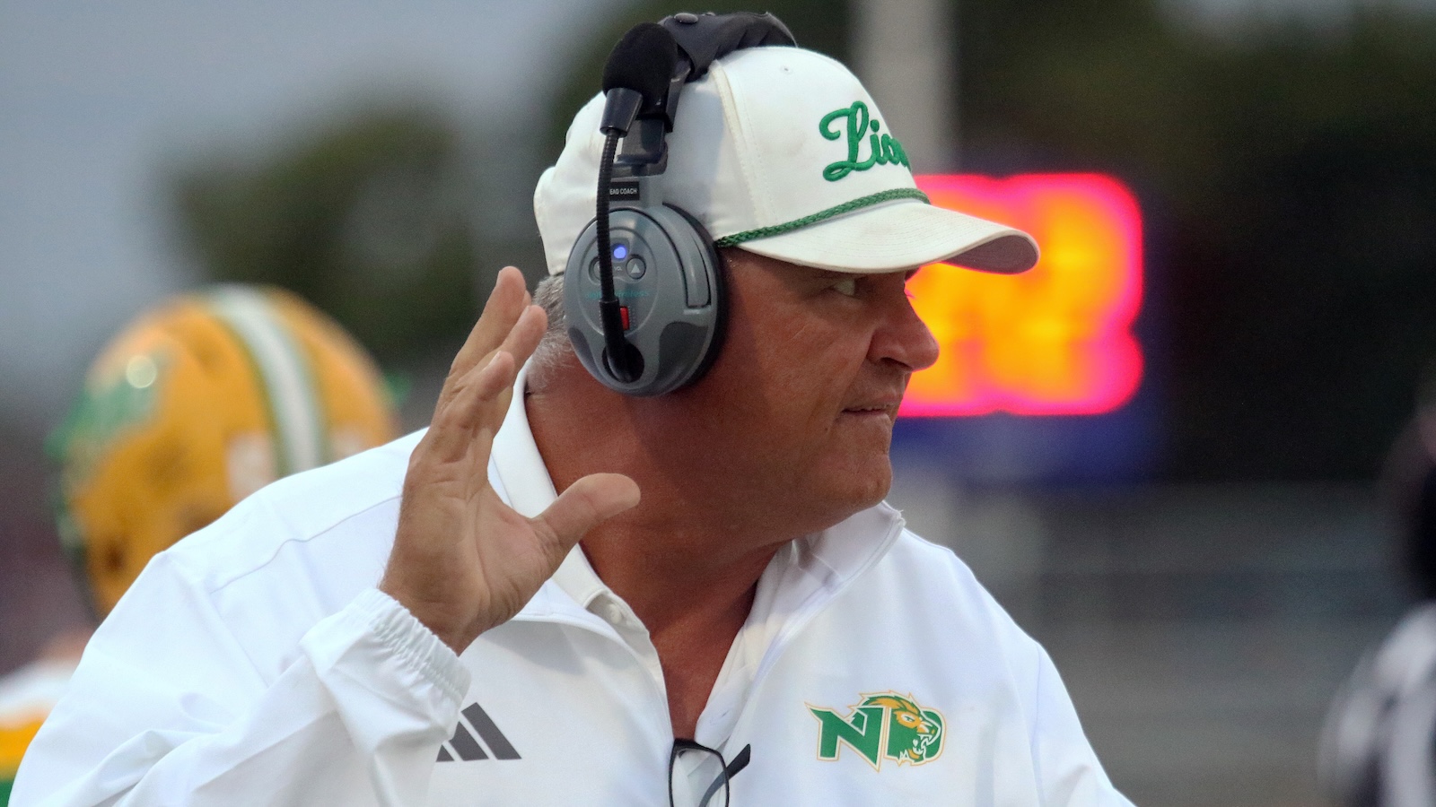 Football coach on the sidelines wearing a white cap and headset, signaling with his hand.