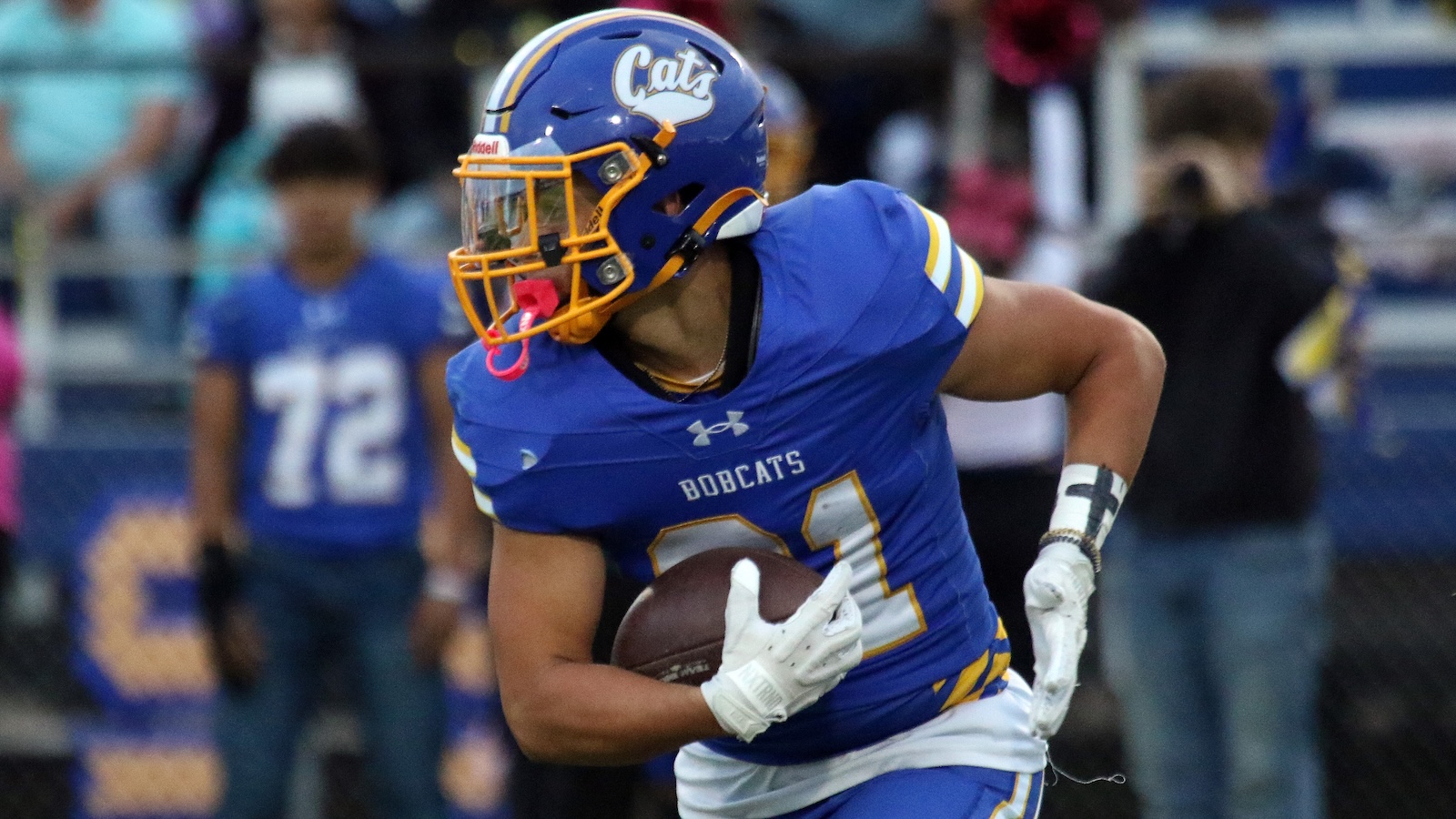 A football player in a blue Bobcats uniform runs with the ball, wearing a blue helmet with a yellow faceguard and'Cats' logo.