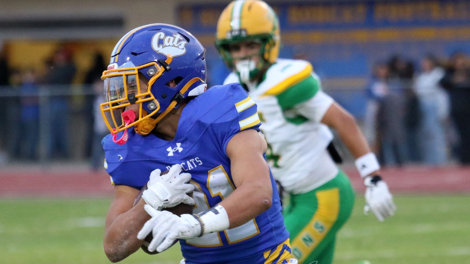 Football player in blue and gold jersey running with the ball, pursued by a defender in green and yellow.