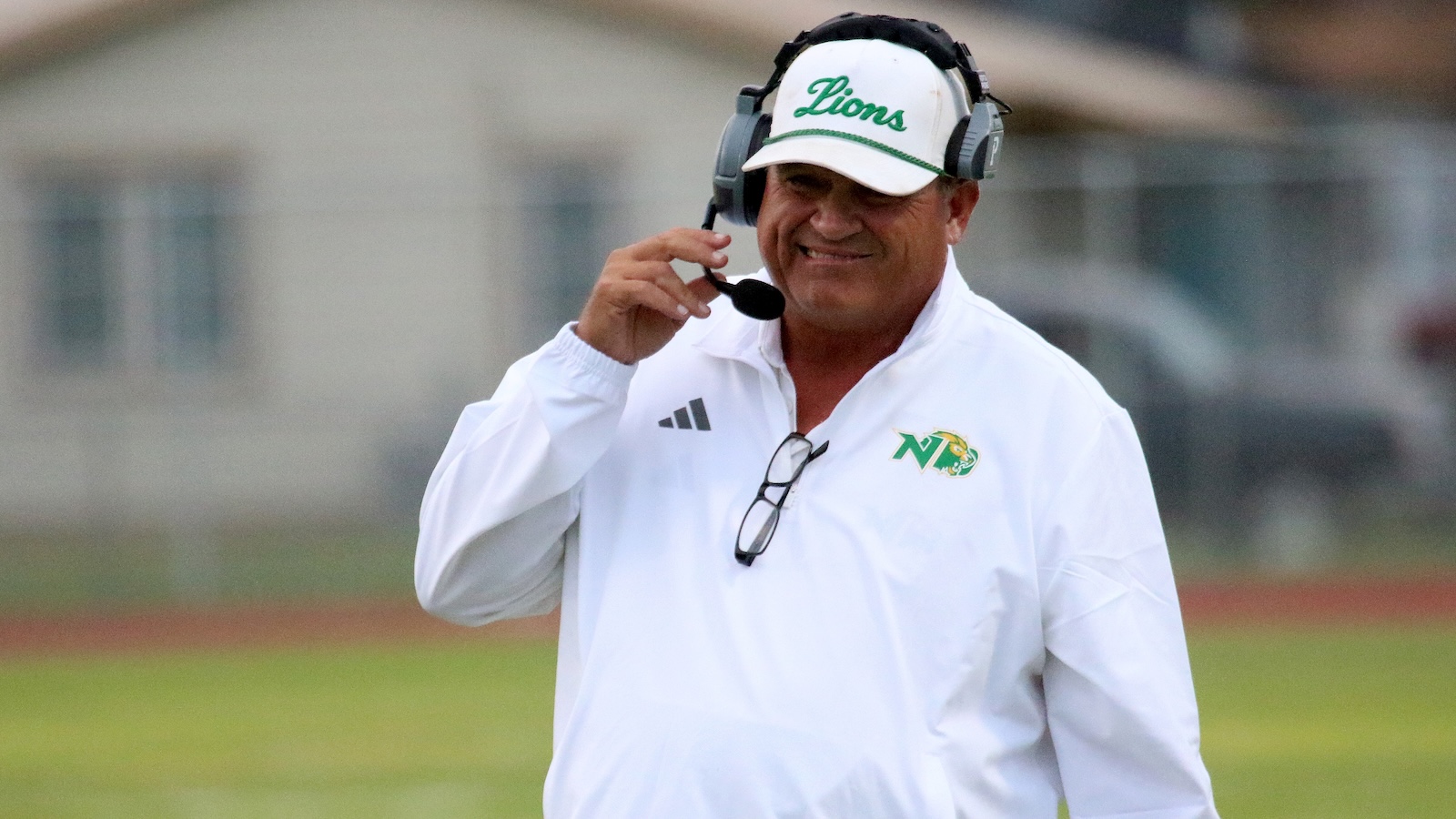 Coach on a field wearing a white pullover and headset, adjusting the microphone in front of a blurred schoolyard backdrop.