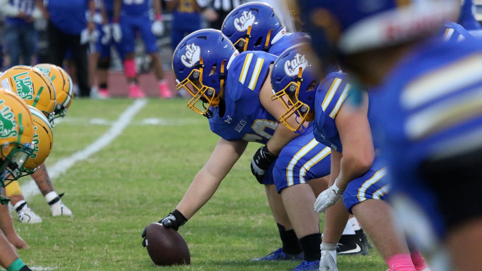 Blue team offensive line crouched at the snap, football on the ground, facing yellow-green defenders.