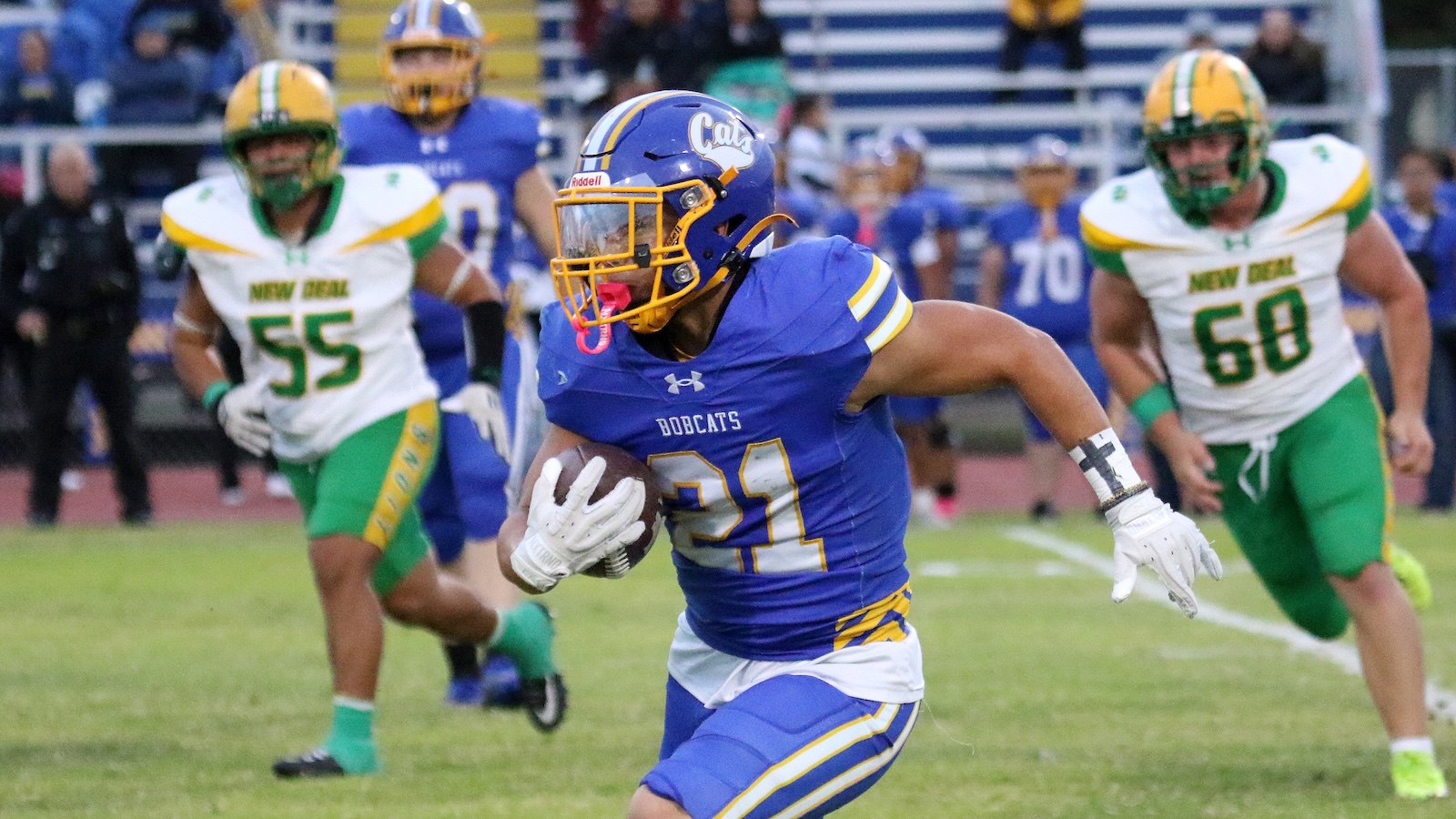 Bobcats football player wearing number 21 in blue and gold runs with the ball, pursued by two green-and-white defenders on a grassy field.