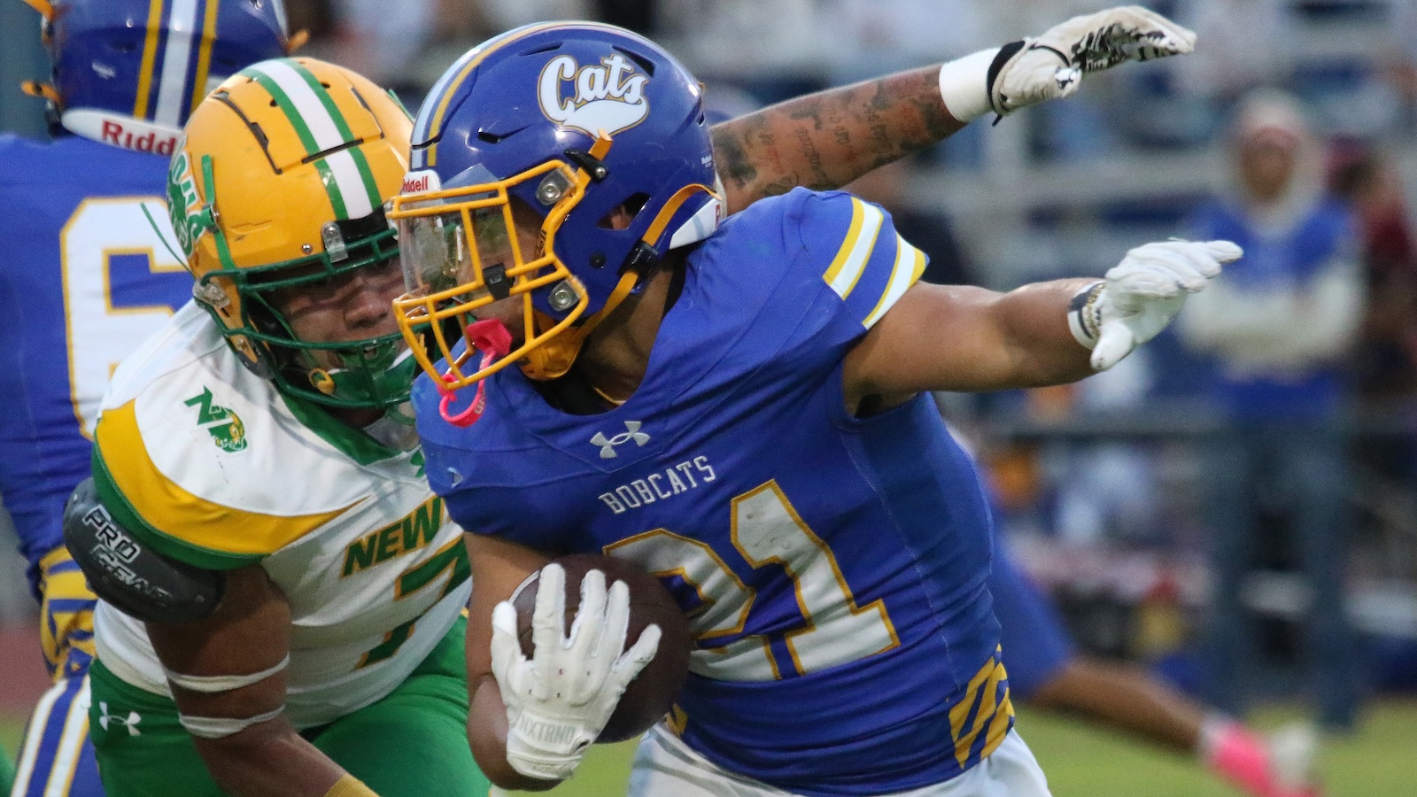 Bobcats running back in blue with the ball tries to break free as a green-and-yellow defender closes in for the tackle.