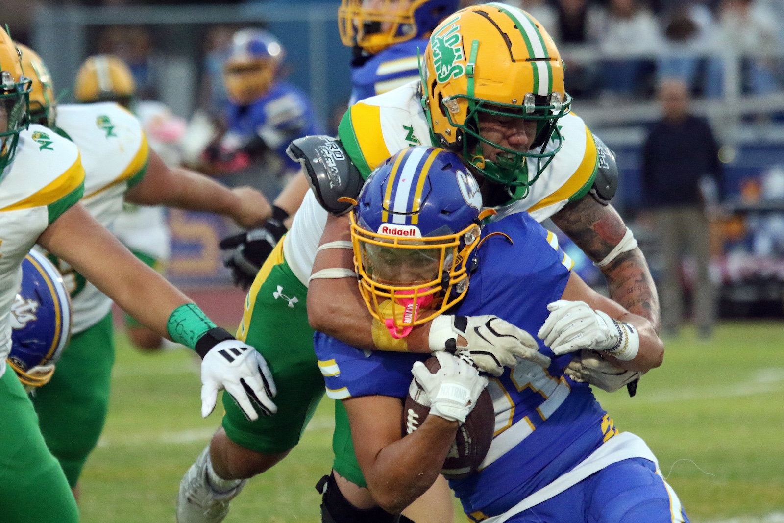 American football play: blue-and-gold team player clutching the ball while tackled by a green-and-yellow defender in a crowded field