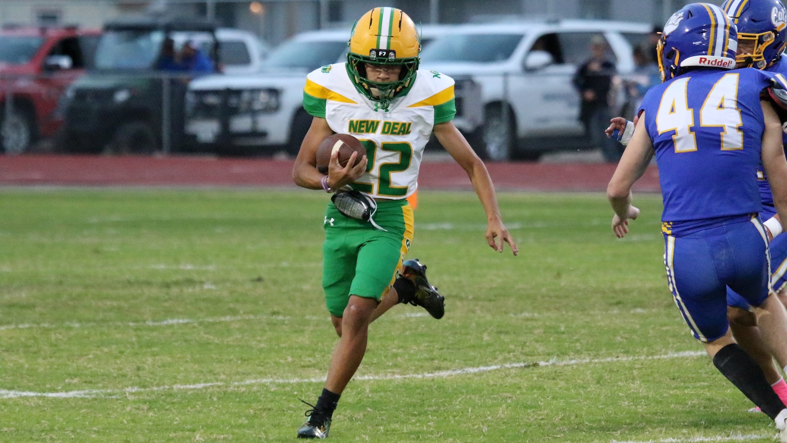 Football player in green and yellow uniform runs with the ball during a game, pursued by blue-clad opponents on a grass field; number 22 visible on jersey.