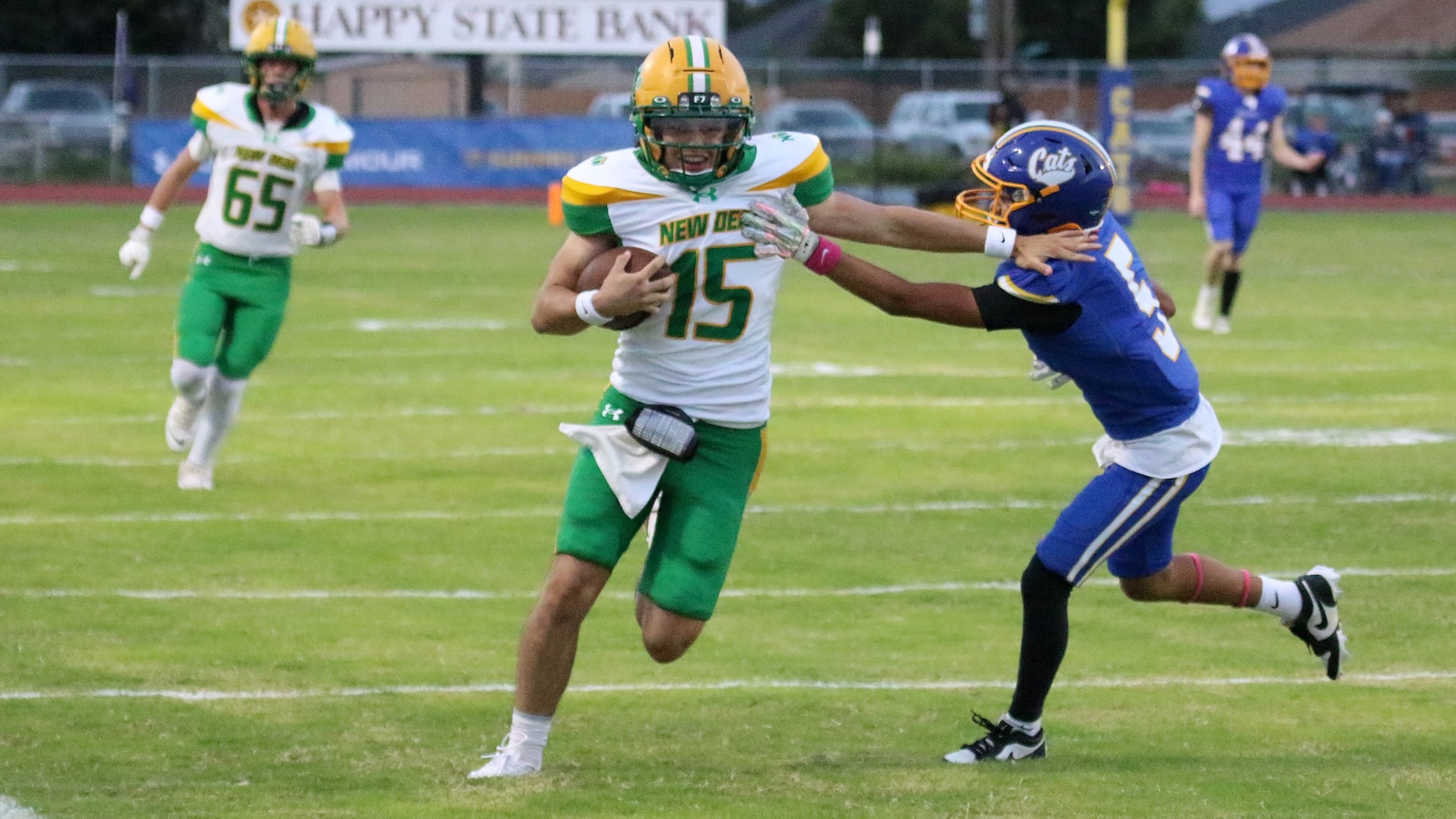 Football player in white and green jersey number 15 runs with the ball as a blue-clad defender reaches in for a tackle on a grassy field.