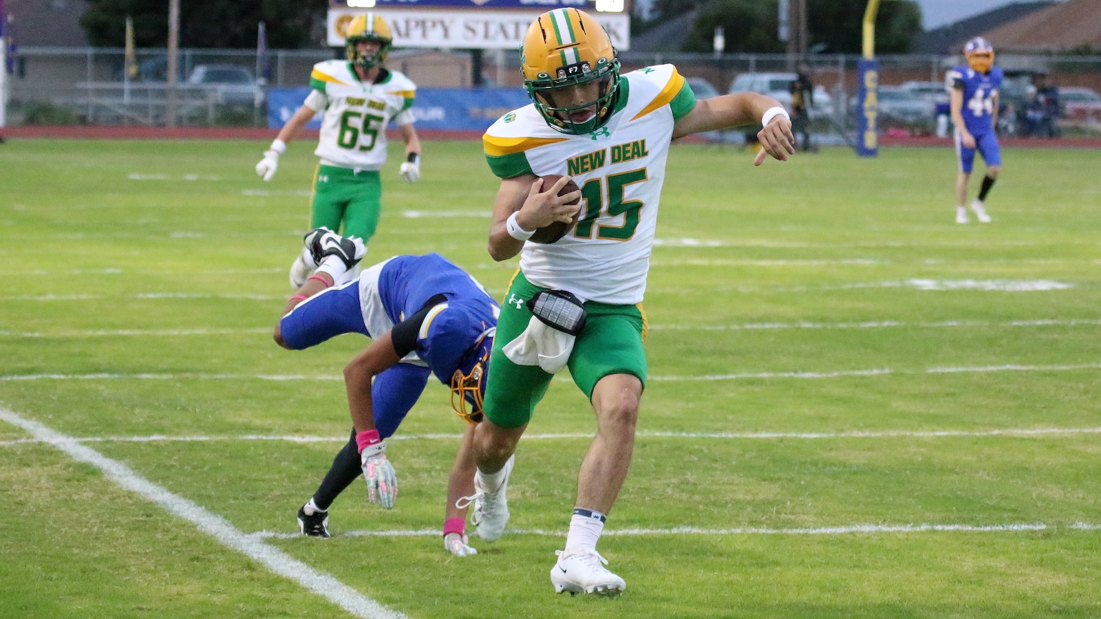 Football player in a New Deal white-green jersey (number 15) runs with the ball while a blue-clad defender dives behind him on a grassy field.