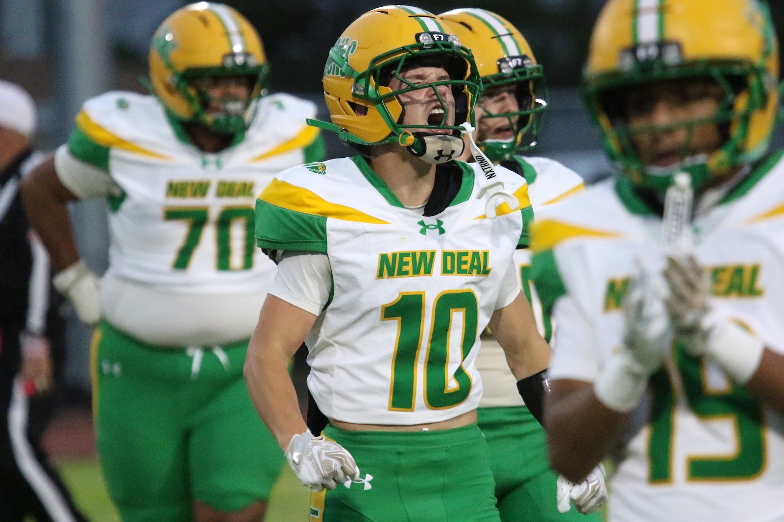 New Deal football player wearing #10 jersey shouts with teammates in green and yellow uniforms behind him on the field, mouthguard visible.