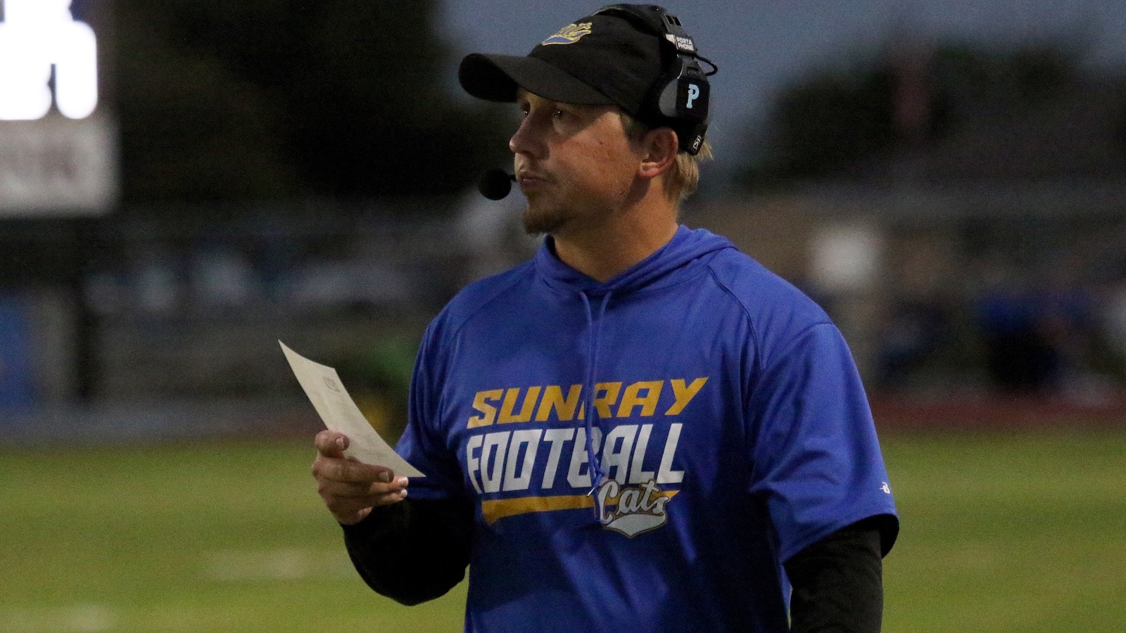 Side-profile of a football coach wearing a blue SunRay Football Cats shirt, headset mic, and holding a play sheet on the field.