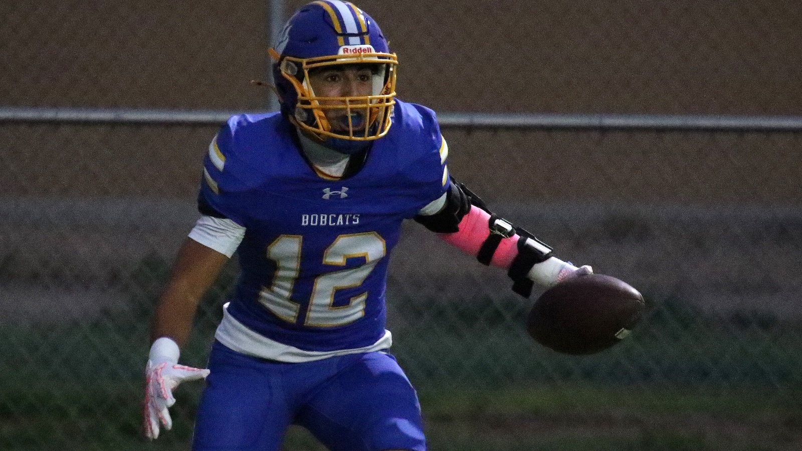 Football player in blue and gold Bobcats uniform holding a football, wearing a gold helmet and pink armband, running on a field.
