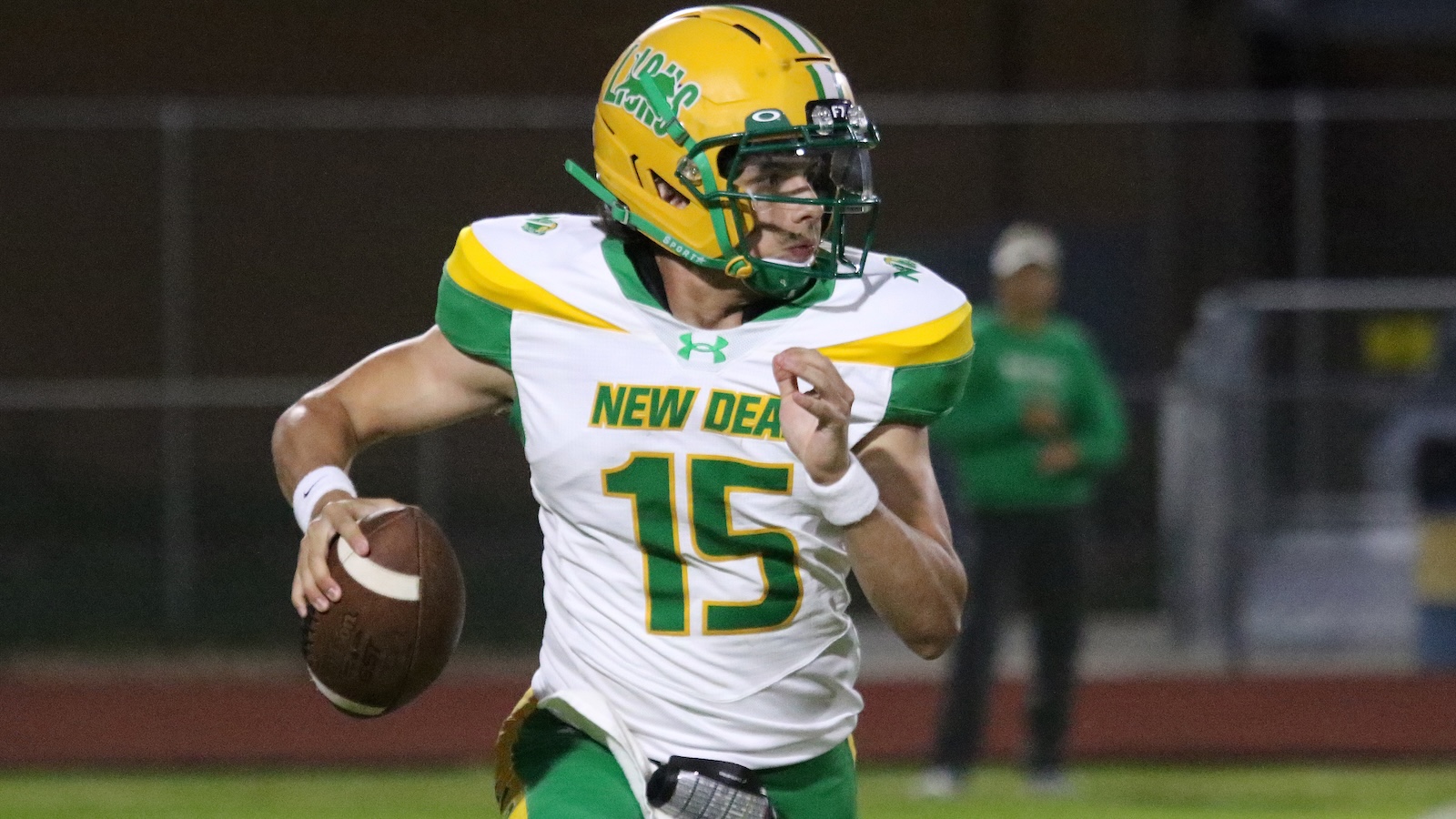 New Deal quarterback wearing number 15, in white/green/yellow, cradling the football and looking downfield during a night game.