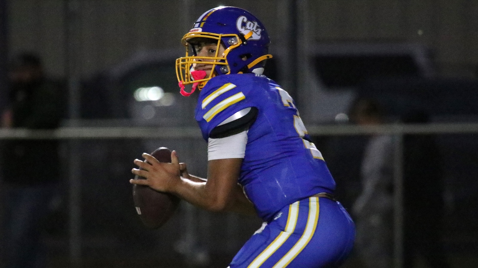 American football player in blue and yellow uniform clutching a football, ready to throw or run during a night game.