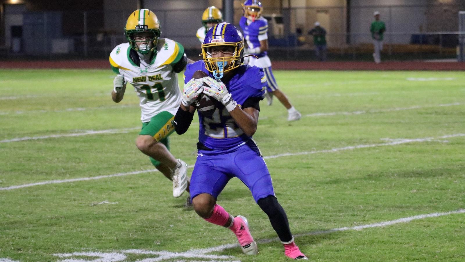 A football player in a purple uniform and yellow helmet runs with the ball while being chased by a defender in a green and white uniform on a grass field at night.