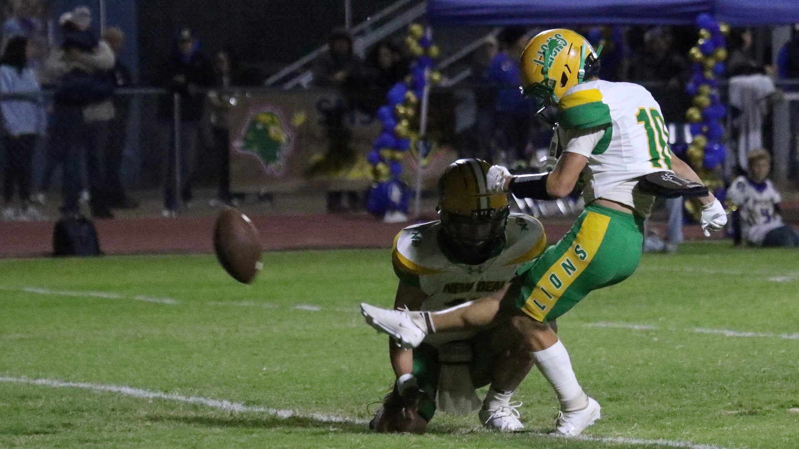 Two football players collide on a grassy field during a night game as the ball spins in the air nearby.