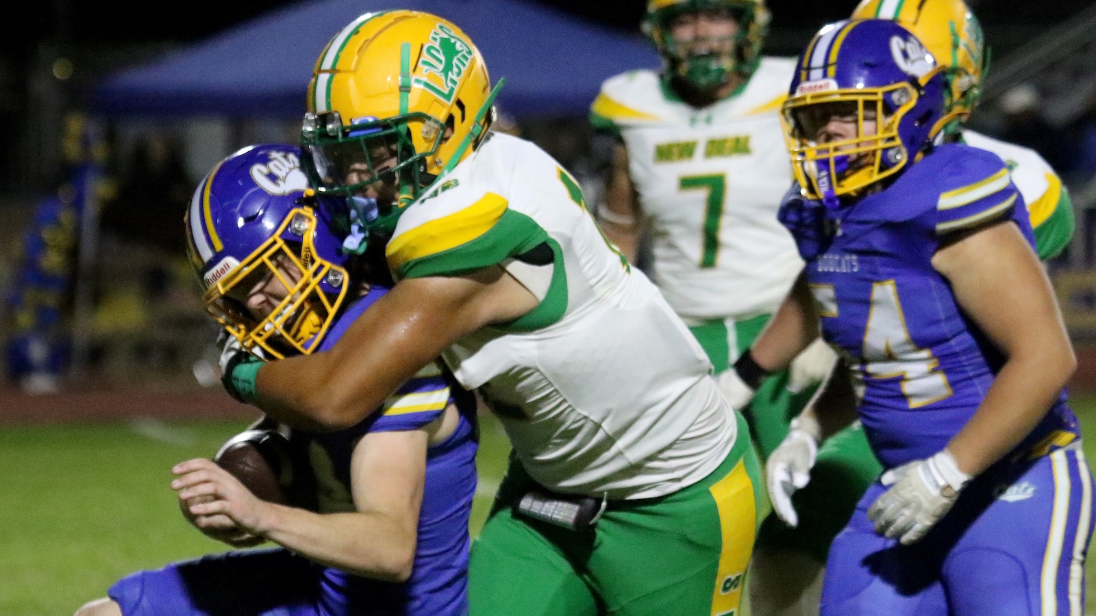 Football players collide as a green-and-white defender tackles a purple-and-gold opponent during a night game.