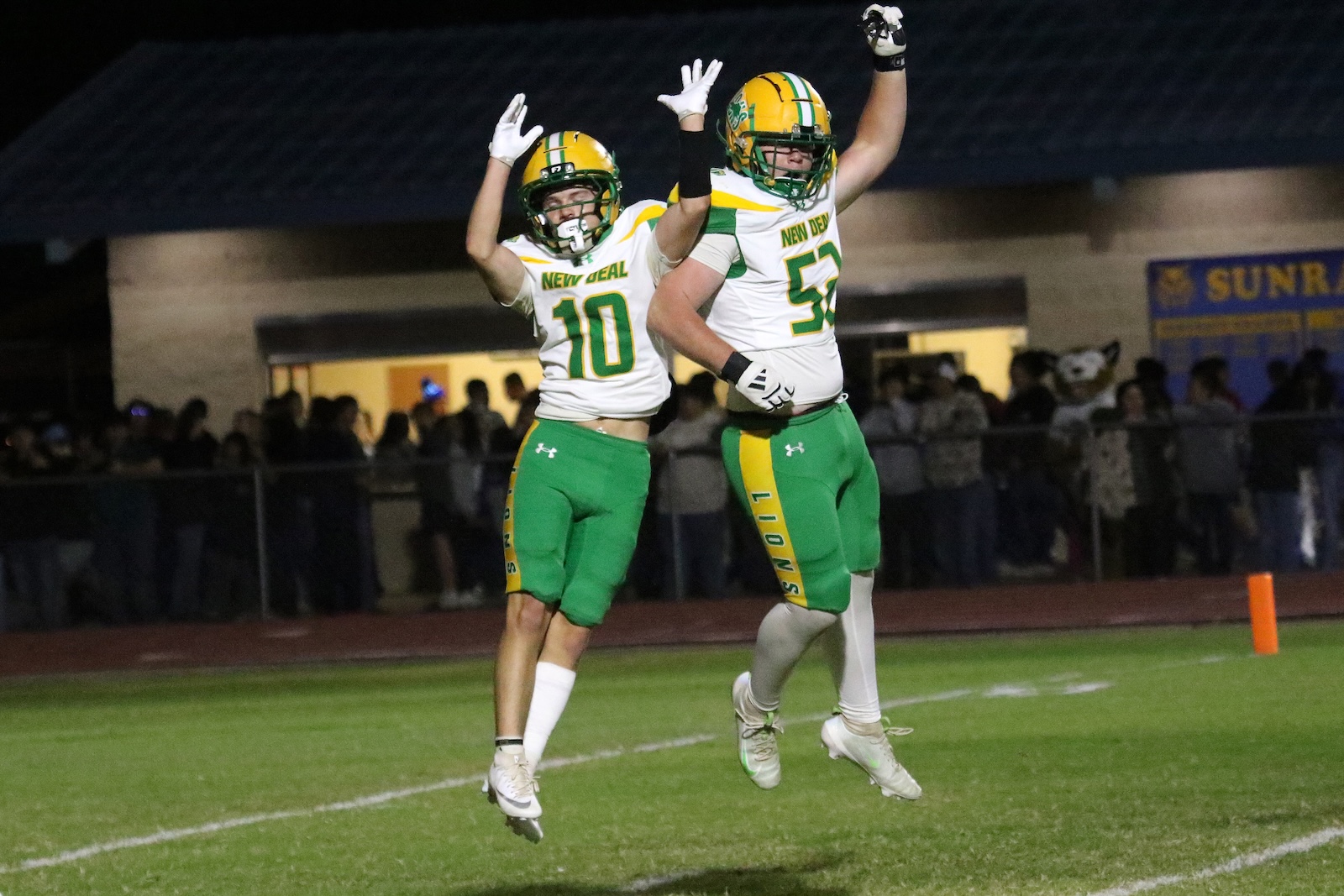 Two football players in green and yellow uniforms jump in celebration on a night game, arms raised, with a cheering crowd in the background.
