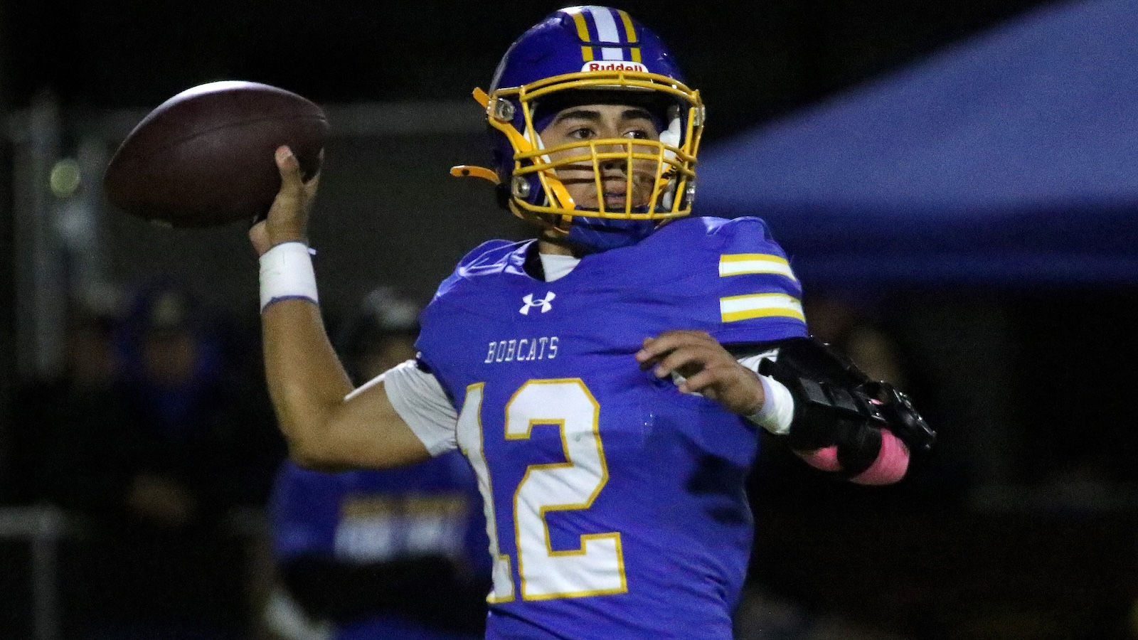 Quarterback in blue and gold Bobcats uniform throwing a football at night.